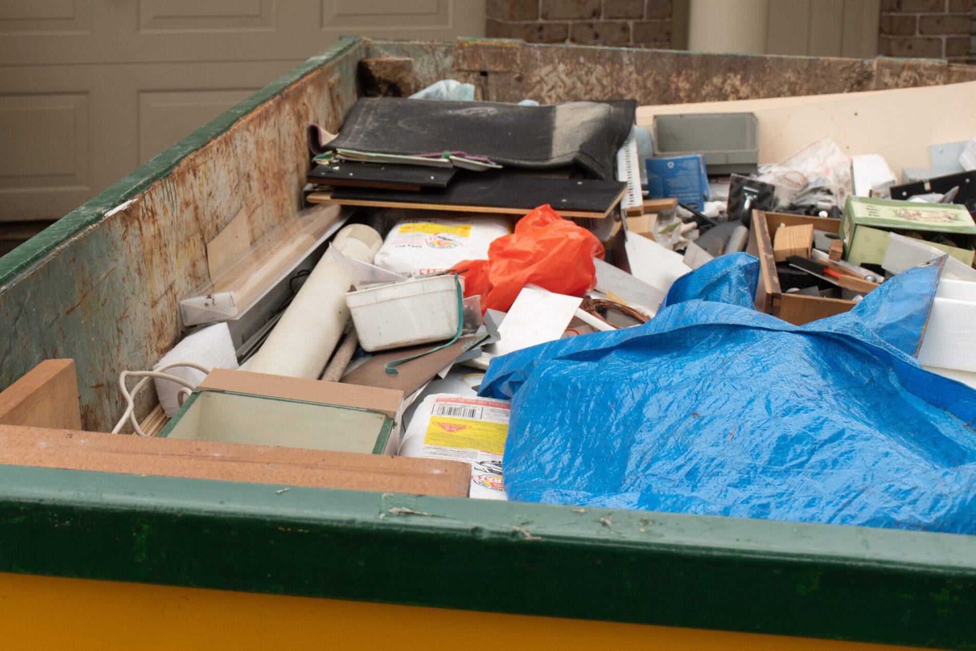 The image shows a large, rusted dumpster filled with various discarded items. It seems to be used for a clean-out or a large disposal project. Visible items inside the dumpster include old electronic equipment, such as a computer monitor, various papers and folders, pieces of wood or frames, what appears to be rolled up posters or plans, and a lot of miscellaneous debris. Part of the debris is covered with a blue tarpaulin, which might be there to protect some of the contents from the weather or just discarded along with the rest of the items. In the background, there is a closed garage door, suggesting that the dumpster is placed in a residential or commercial area, possibly for renovation or moving out purposes.