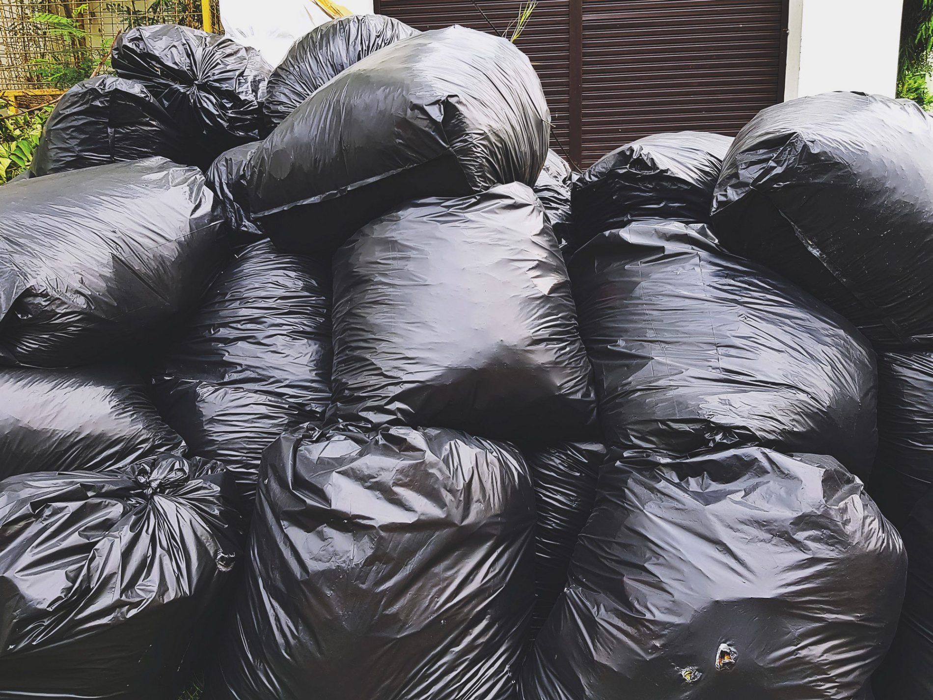 The image shows a large pile of filled black rubbish bags. It looks like they are stacked up for disposal or pickup. The background includes part of a building with a closed window or shutter, and there's some vegetation indicating the bags are likely outside. The content of the bags is not visible, but they are typically used for holding waste or discarded items.