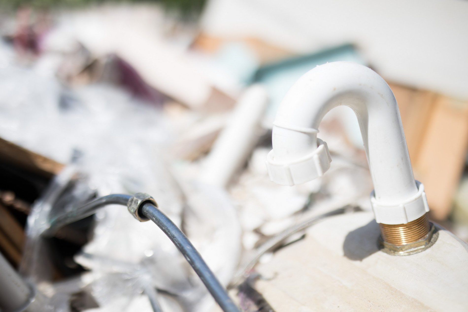 The image shows a close-up of a white plastic P-trap connected to what appears to be a sink's drain, typically used in plumbing to prevent sewer gases from entering a building. The backdrop is blurred, but it seems to be a cluttered area with various discarded items or debris, indicating the setting might be a place where plumbing work is being done or materials are being discarded. The P-trap is in focus, and the sunlight suggests an outdoor environment or a well-lit indoor workspace.