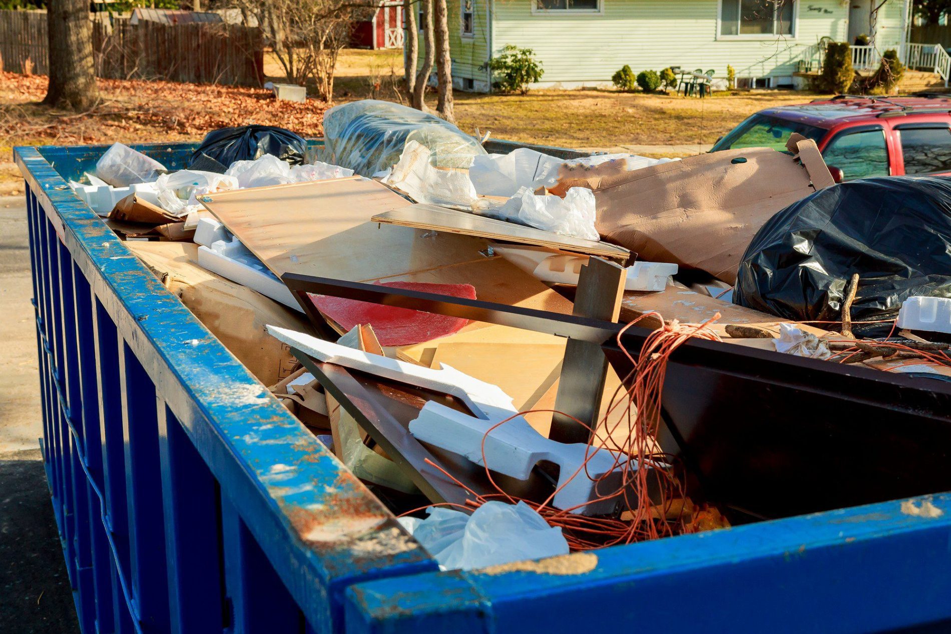 The image shows a blue dumpster filled with various types of debris and discarded items. It appears to be overloaded with materials such as wood panels, plastic bags, pieces of foam, and other miscellaneous waste, indicating that there could have been a recent clean-out or construction-related activity. In the background, there's a quiet residential area with houses and parked cars, suggesting that the dumpster is located near a residential neighborhood. The lighting in the photo suggests it could be either morning or late afternoon.