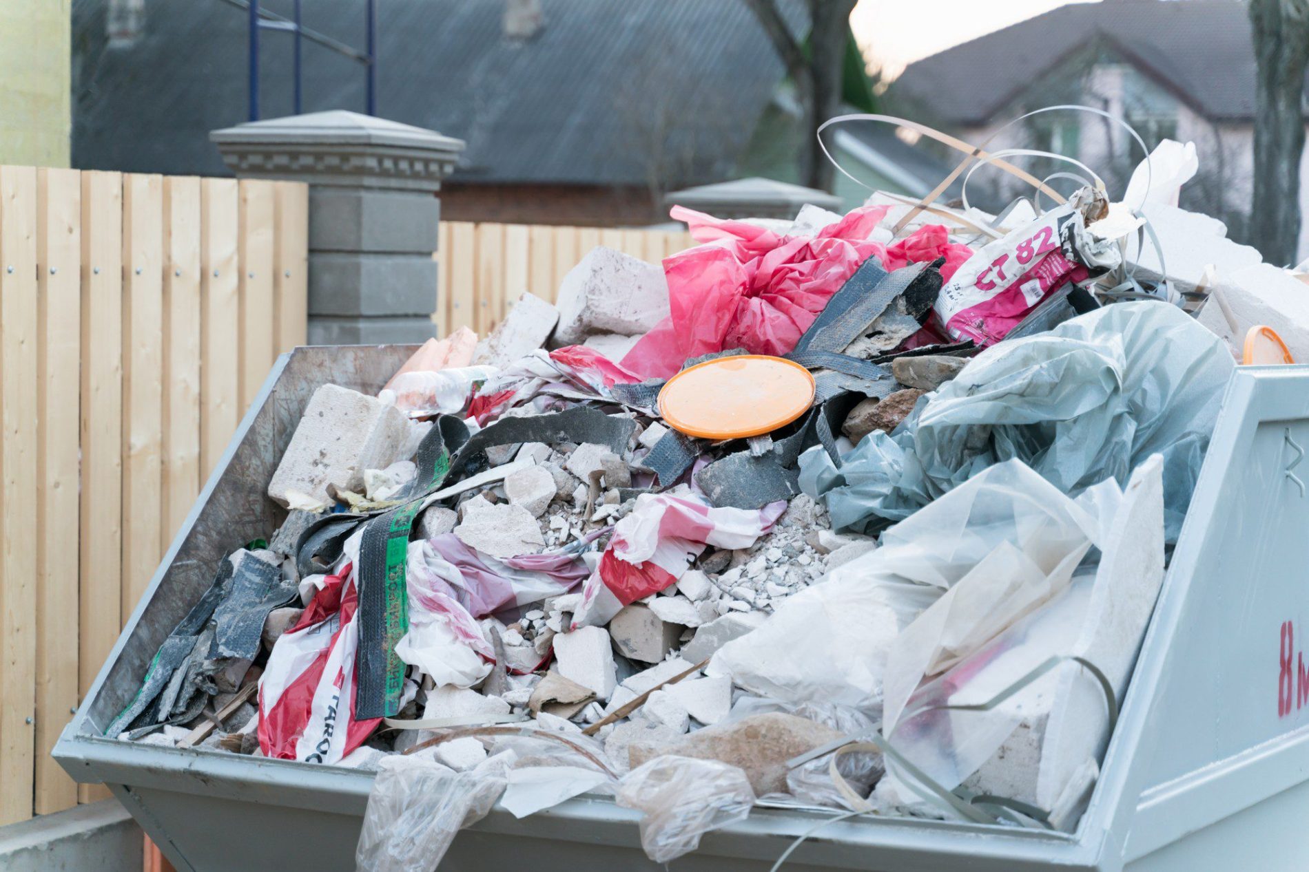 The image shows a large dumpster filled with various types of debris and waste materials. There is a mix of construction debris, including pieces of broken concrete, fragments of material that could be drywall or plaster, pieces of wood, a section of metal rod, and various types of plastic such as a plastic sheet or tarpaulin. We can also see thin strips of plastic that might be from plastic bags, some fabric, and a large orange lid, potentially from a paint bucket or other container. The dumpster is situated in front of a fence, and there are buildings and trees in the background, indicating the setting might be a residential area or a place undergoing renovation or construction.