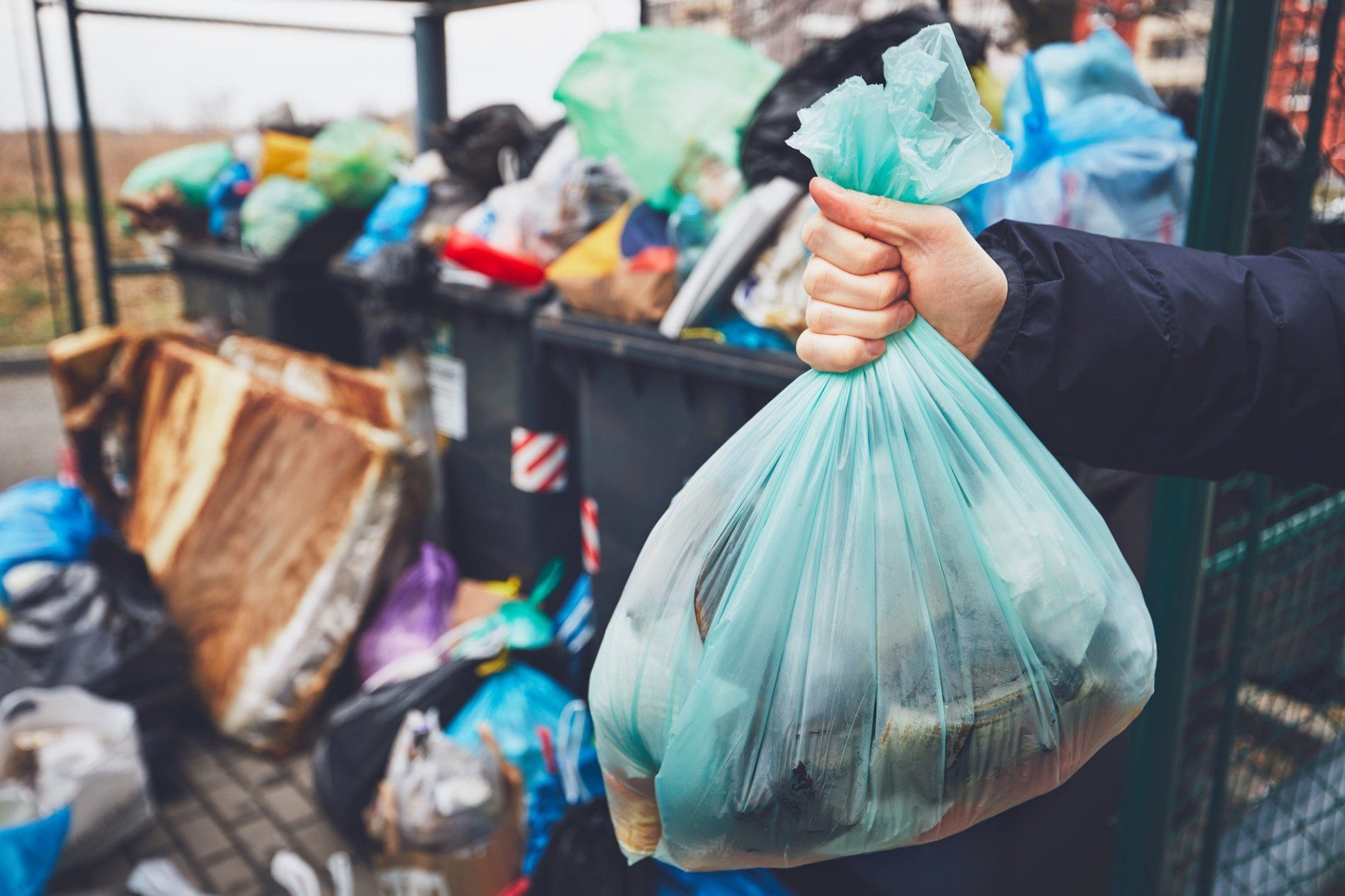 The image shows a person's hand holding a tied light blue plastic rubbish bag. In the background, there are multiple bins overflowing with trash. The bins are filled with various types of waste, including plastics, cardboard, and other materials. The cluttered and overcrowded state of the bins suggests that it may be a communal waste disposal area, possibly in a residential area. The focus is primarily on the hand holding the rubbish bag, indicating the action of disposing waste.