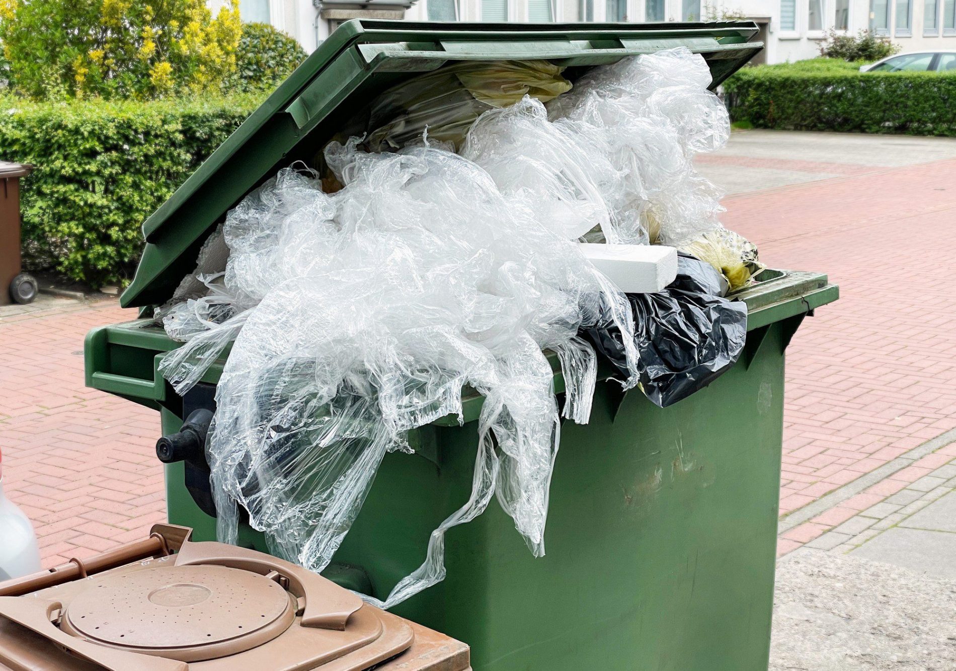 The image shows an overflowing green waste bin. It's filled with various types of trash, most prominently a large amount of clear plastic wrapping material which is spilling out of the bin. There are also other items of waste such as foam containers and black plastic bags visible, suggesting that the bin has reached its capacity and can no longer be properly closed. This could lead to potential littering issues if the waste is not managed soon. The bin is situated on a brick surface, and there is some vegetation and a building in the background.