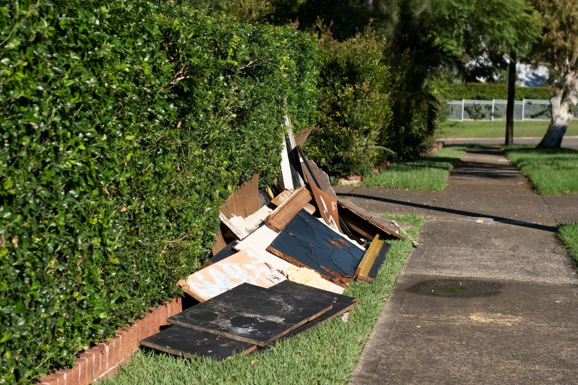 The image shows a pile of discarded items, which appear to be pieces of broken furniture and wood, placed on a grass verge next to a sidewalk. The items are likely set out for bulk waste collection. In the background, there is a nicely trimmed hedge running parallel to the sidewalk, and a street is partially visible. The overall setting suggests a residential area on a sunny day.