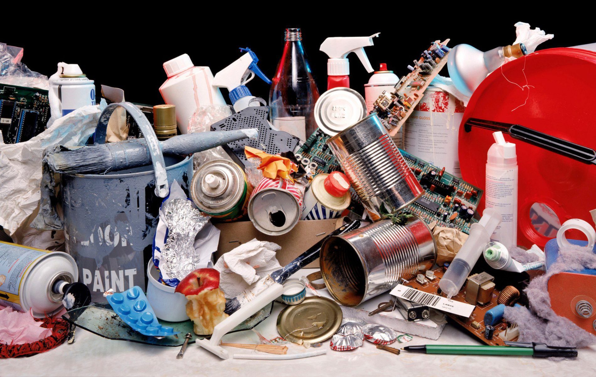 The image shows a collection of various discarded items, suggesting a heap of waste or rubbish. Included are:

- Plastic containers and bottles, some with product labels
- Empty food cans and a dirty aluminium foil piece
- A half-eaten apple
- Electronic waste, such as parts of a circuit board
- Used paint cans and a paint roller with blue paint
- A plastic bucket with the label "PAINT"
- Miscellaneous items like a spoon, a pen, a light bulb, and a small shovel
- Packaging materials, including torn plastic bags and paper
- A red plastic lid and a spray cleaner bottle with a trigger handle

This assortment represents typical refuse, pointing to issues of consumption, waste management, and environmental pollution.