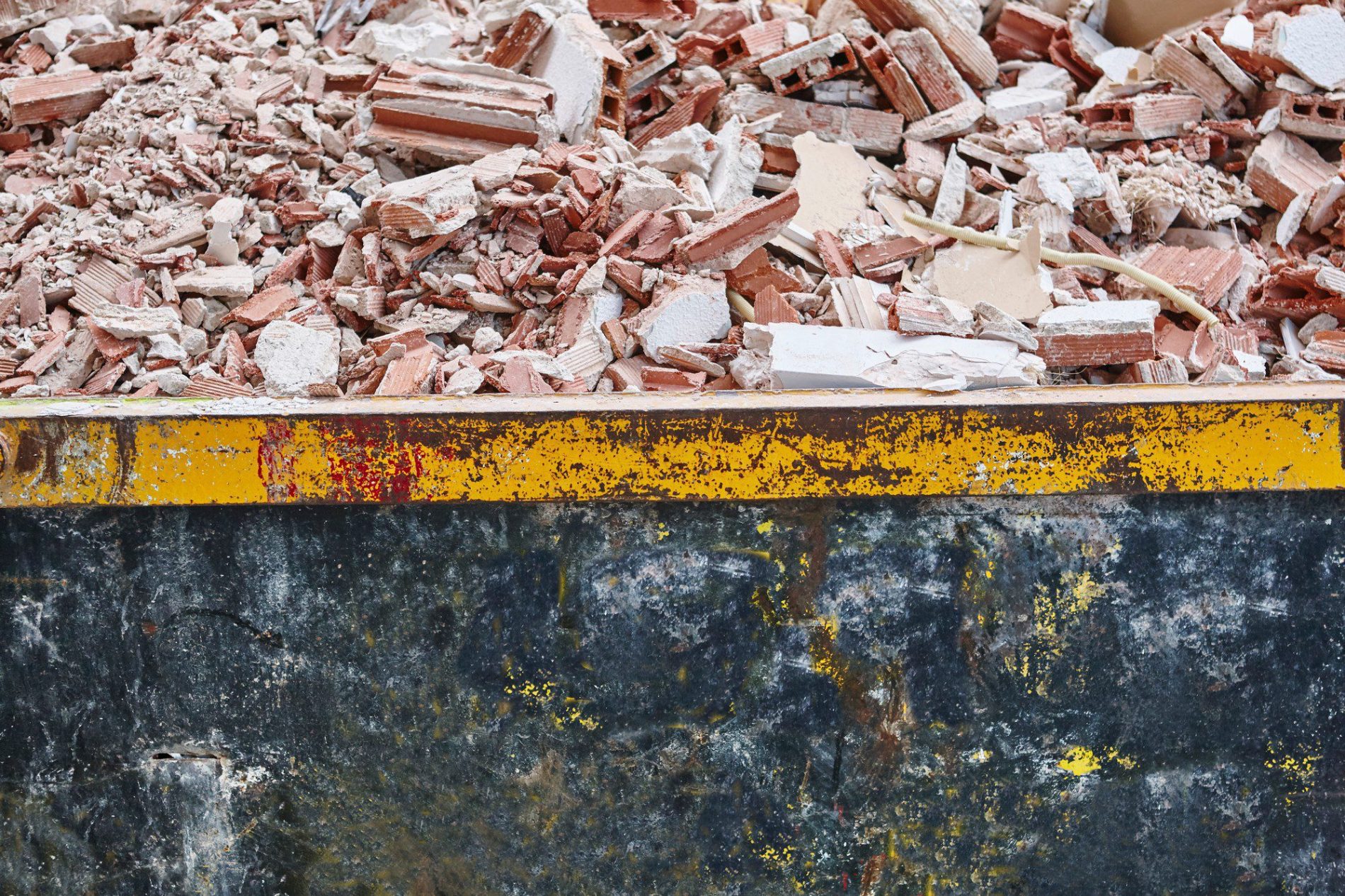 The image shows a large container filled with construction debris, primarily consisting of broken bricks, chunks of concrete, and pieces of plaster. The container itself has a visible yellow stripe along the top edge with some red and yellow paint flaking off, indicating wear and exposure to the elements. This sort of container is typically used on construction or demolition sites to collect and transport waste materials for disposal or recycling.